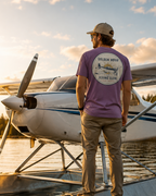 A man wearing a purple t-shirt with a 'Golden Hour Flying Club' graphic stands on the floatplane's pontoon, facing a white and blue Cessna seaplane on a lake at sunset.